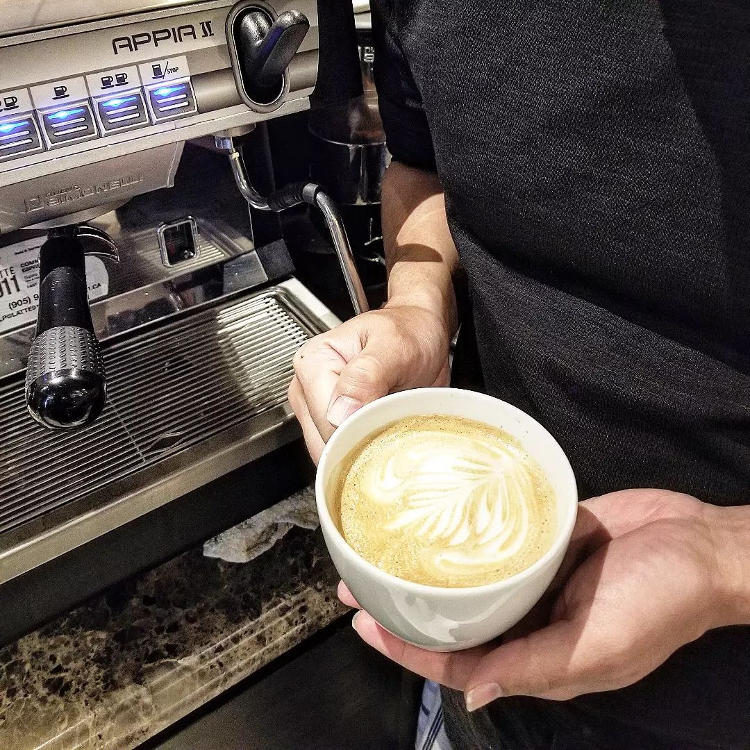 Latte with leaf on the top in a ceramic cup, with a barista machine in the background and person standing beside holding the mug.