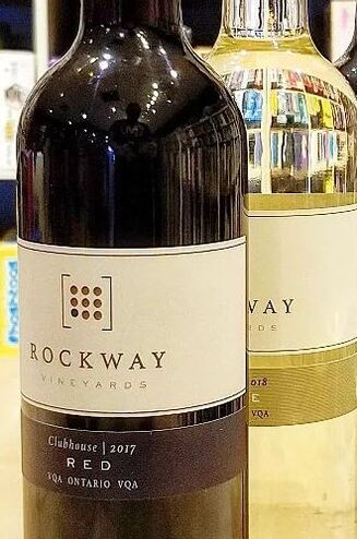Two glass bottles, standing upright on a table. The front bottle is Red wine by Rockway Vineyards and behind it is a white wine from Rockway Vineyards. They are standing on a wooden table with games reflecting through the glass. 