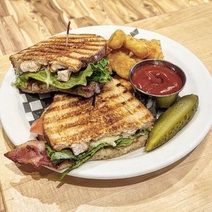 A Chicken sandwich with grilled lines that includes bacon, lettuce and sauce, tater tots beside a small metal cup of ketchup and a slice of pickle on a ceramic plate. It sits on a table with board games and a wooden floor featured in the background.